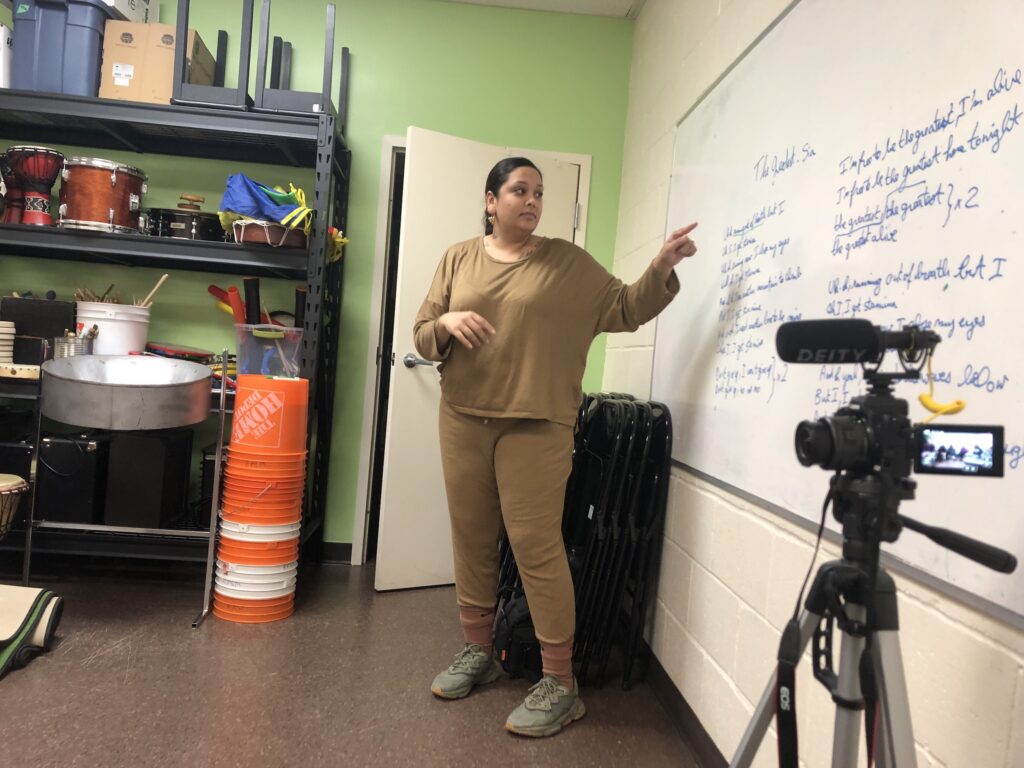 Gaitrie Persaud standing in front of a white board, showing words of the lyrics The Greatest by Sia.