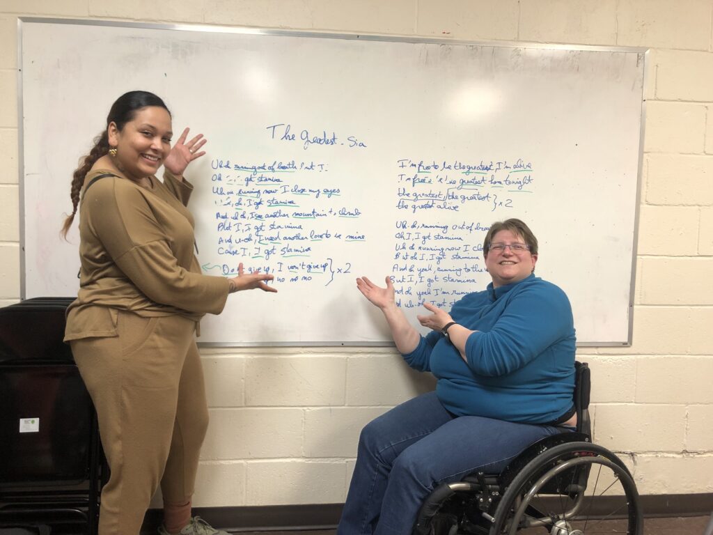 Gaitire standing on the left and Diane sitting in her wheelchair on the right, showing the lyrics of the song The Greatest by Sia.