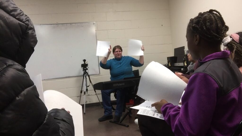 Diane in front of students, sitting in her wheelchair and showing the Braille score