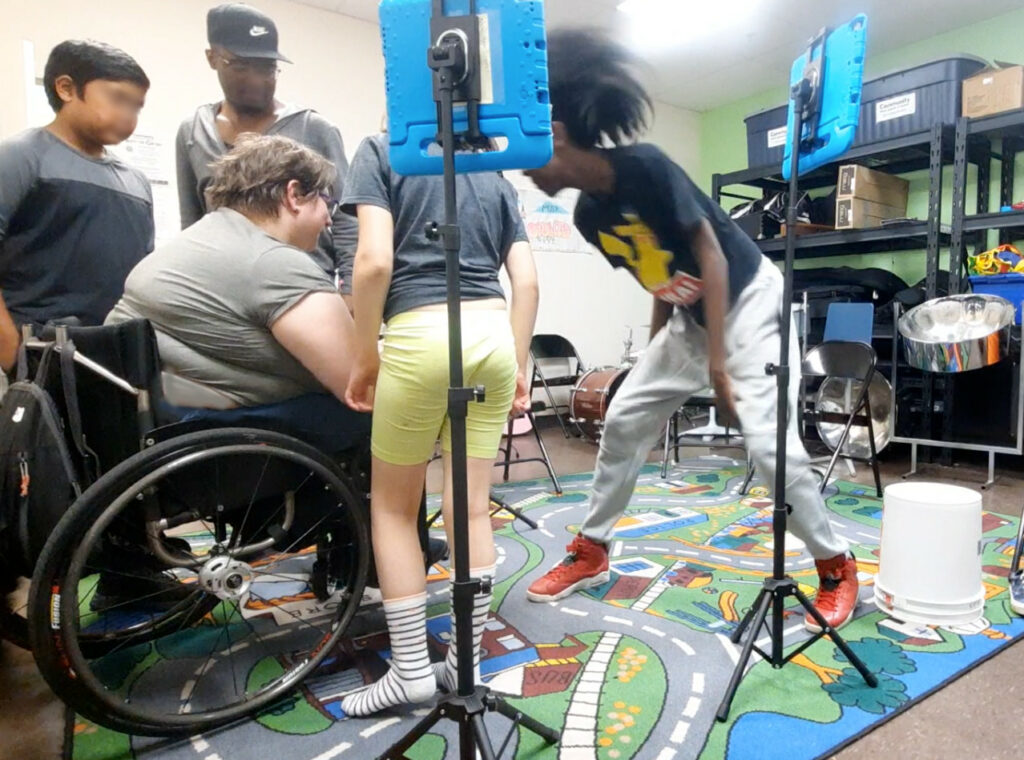 A group of students playing instruments differently in the music room, surrounded with a few iPads on stands.