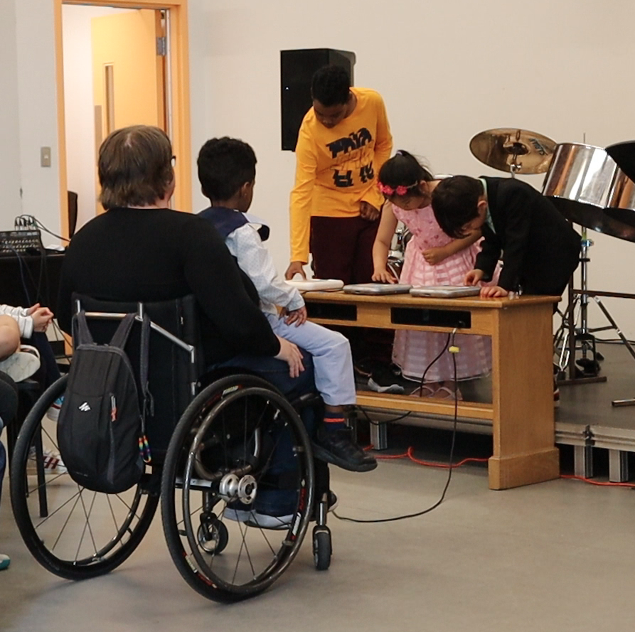 Group of students performing on the Tenori-On. Diane is on the front helping them, sitting in her wheelchair. A little boy sits on her knees.