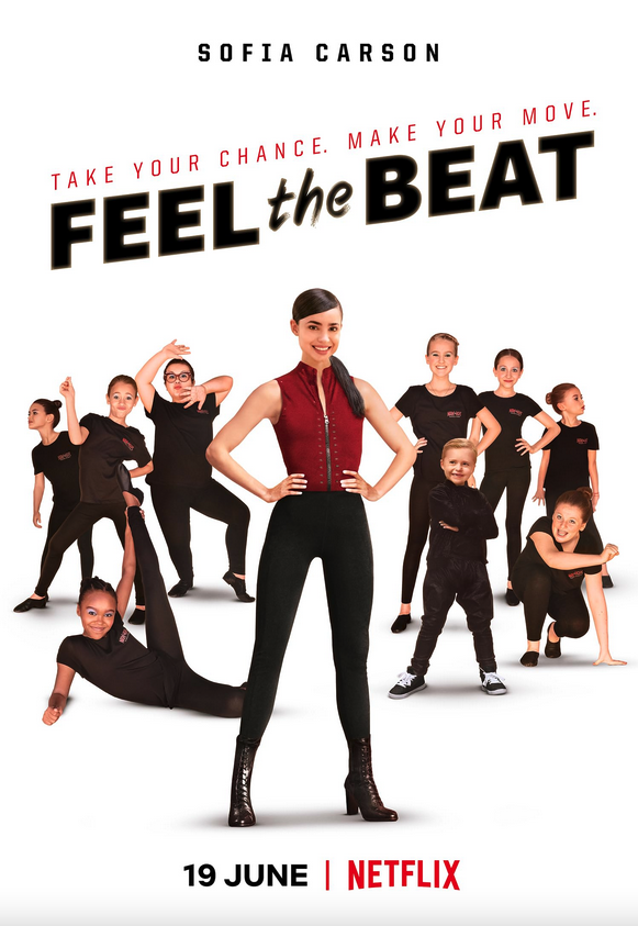 A group of children and teenagers wearing black clothes, holding diverse dancing pauses. A young woman wearing a red top stands in the middle with her hands on her hips, smiling.