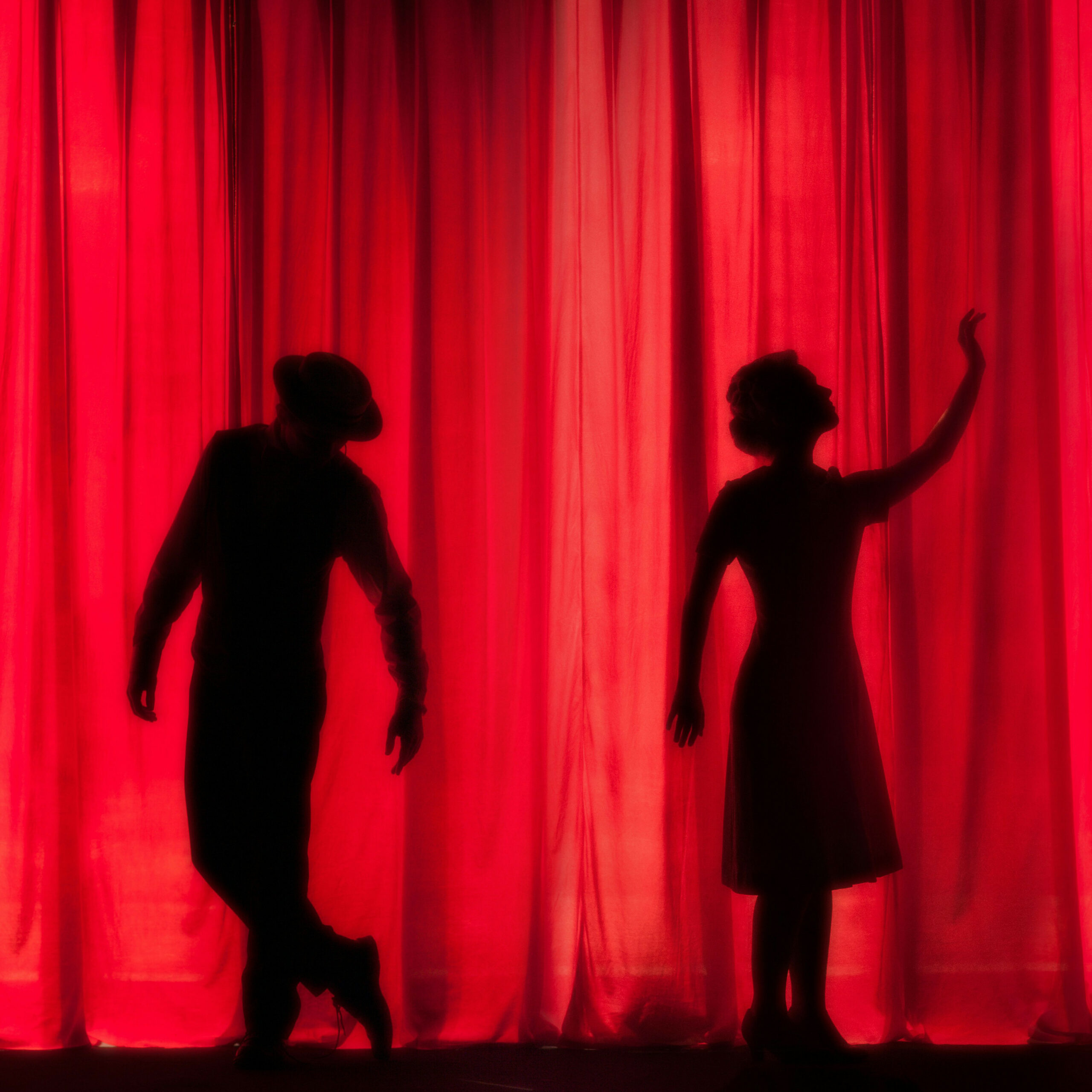 Shadows of two actors in front of a red curtain on a stage. The man on the left is dancing, the woman on the right waves to someone backstage.
