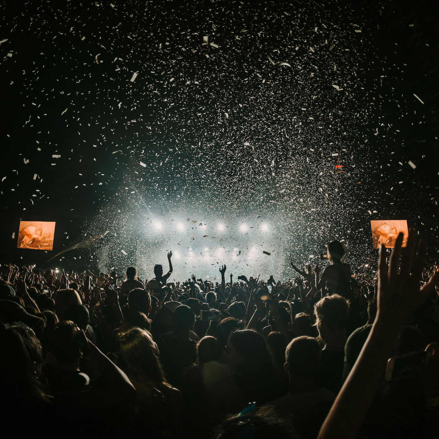 A crowd cheering and throwing confetti. An illuminated stage is in the background, with two orange giant screens on both sides.