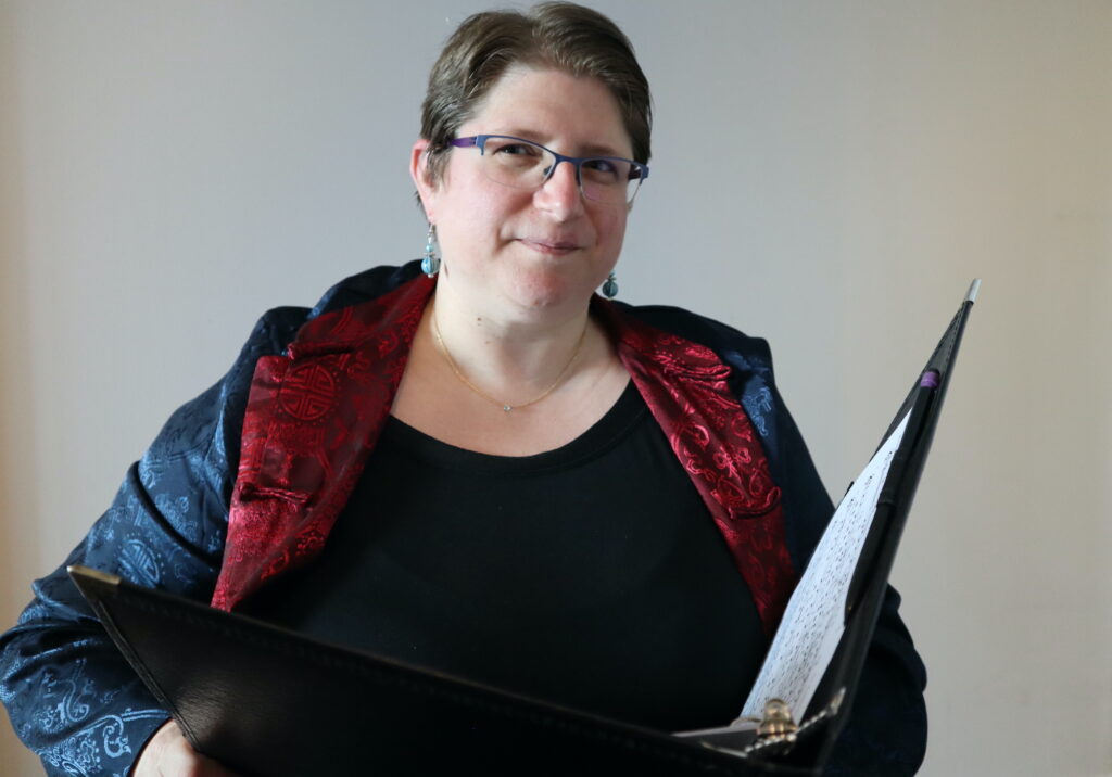 A white woman with short hair and glasses wearing a Chinese outfit and holding a music folder.