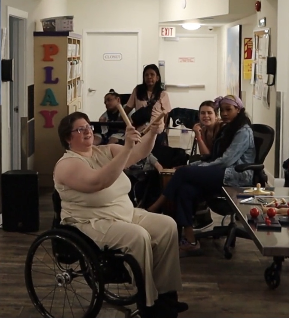 Diane Kolin sitting in her wheelchair, holding sticks to demonstrate a rhythm during the workshop at Safehaven.