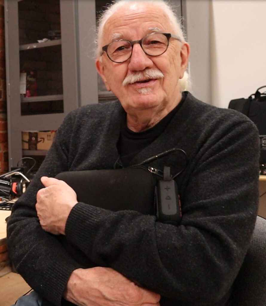 Un homme blanc avec une moustache blanche et des lunettes à monture noire, portant un pull-over gris foncé. Il tient un coussin noir avec une télécommande attachée sur le côté.