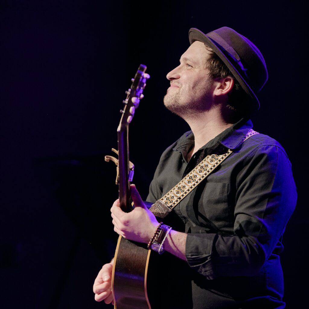 A white man with short brown hair seen from the side, wearing a hat and holding a guitar.