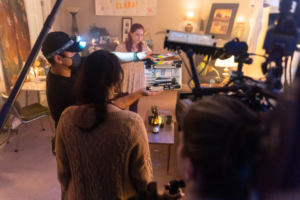A view behind the scene of the movie shooting. Someone holds a clapper on which the text "The sweetest goodbye, Roll A003, scene 2D, take 1" is written. Other people stand behind a microphone boom and a camera.