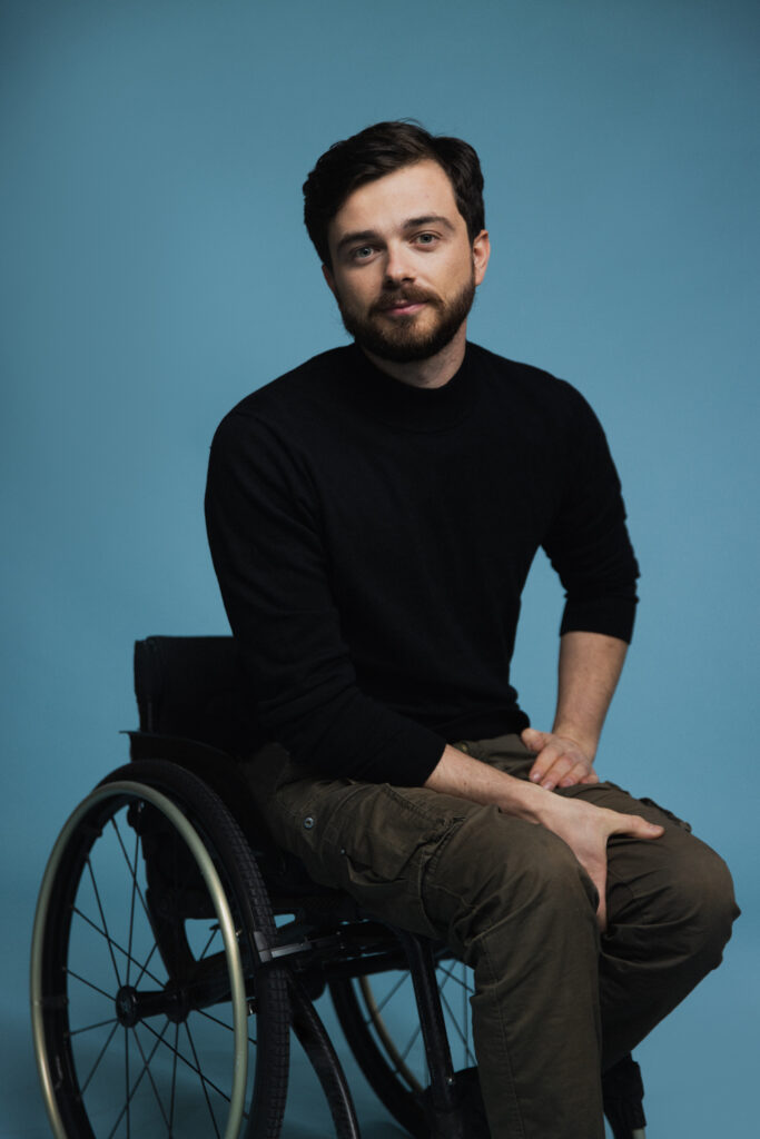 A white man with black hair wearing a black sweater and khaki pants, sitting on his wheelchair. There is a blue background behind him.