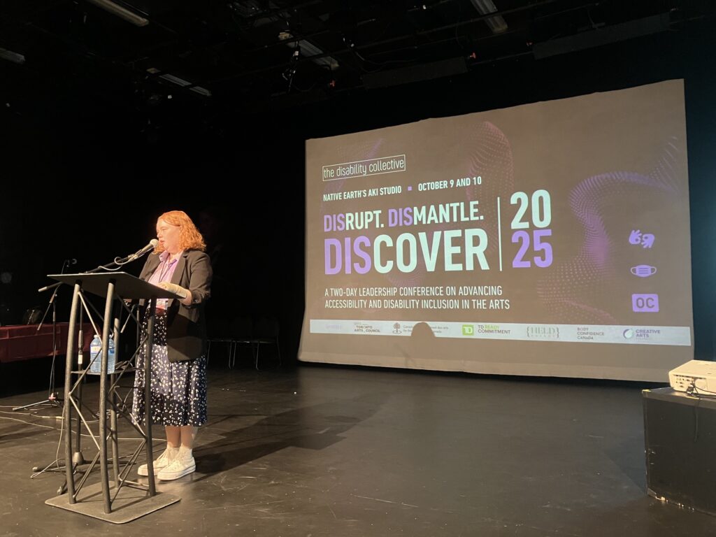 Emily Maxwell, a white woman with curly red hair, wearing a pink top, a black skirt with white dots, and a black vest. She speaks at a stand in a microphone installed in front of her. Behind her is a projected slide showing the poster of the DisCover conference, saying Disrupt, DisMantle, DisCover.