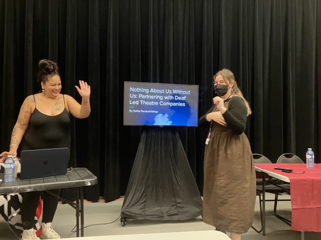 Gaitrie Persaud-Killings on the left, wearing black pants and a black tops, waves at someone. On the right, an interpreter is ready to work with her. A screen shows a slide giving the title of her presentation, Nothing About Us Without Us, Partnering with Deaf Led Theatre Companies.