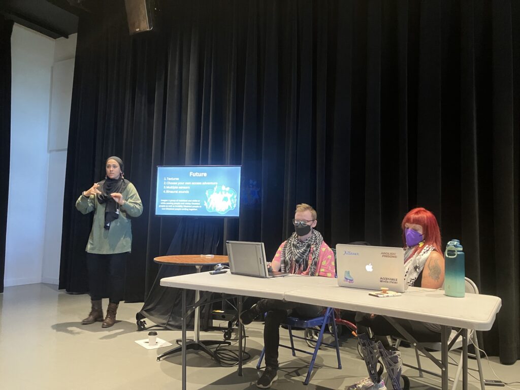 Maxime Beauregard and Erin Ball sit at a table in front of their computers. A screen shows their presentation, the slide says Future and lists what future shows they want to accomplish. A sign language interpreter signs on the left.