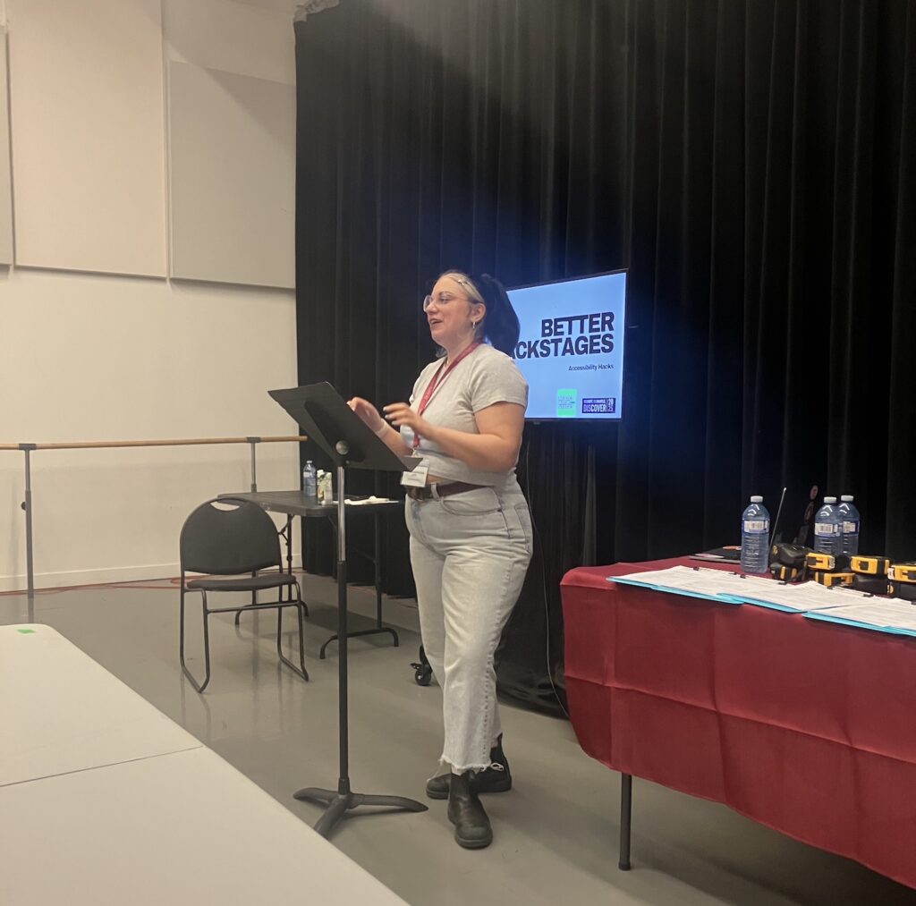 Meghan Sivani-Merrigan, a white woman with glasses and black hair wearing a white t-shirt and light grey pants, standing in front of a stand. On her left, a table with blue clipboards and tape measures.