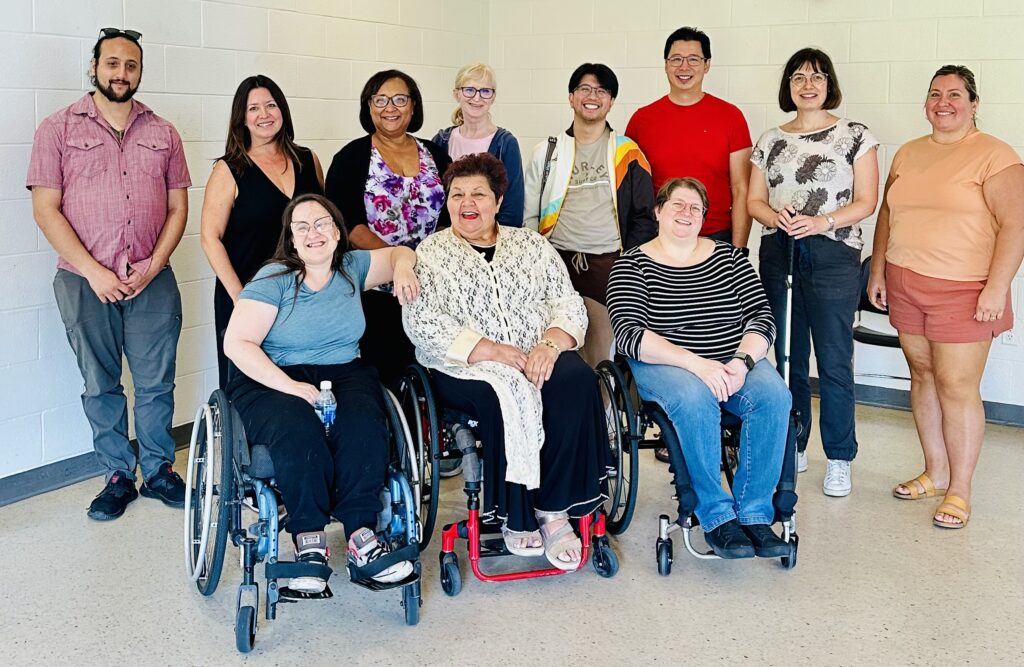 Some members of the group of participants and Diane. Three people sit in wheelchairs, and the rest of the group stands behind. One participant standing on the right holds a white cane.