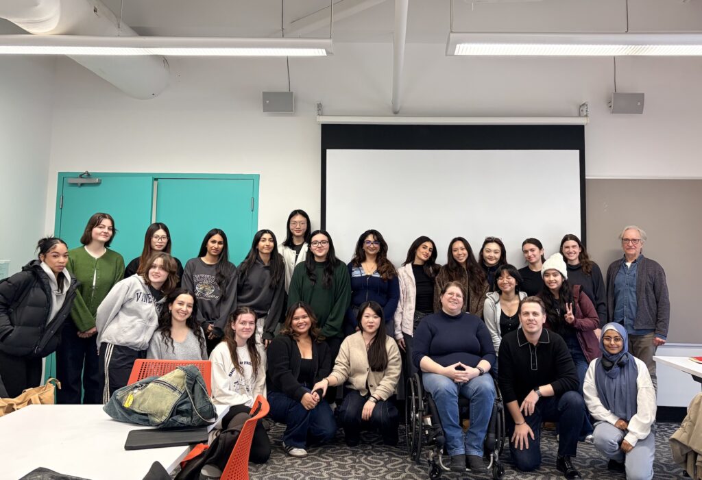 Students of a class in OCAD University of Design. Diane sits in her wheelchair in the first row, Roman Romanov, the teacher of the class, sits close to her, and about 20 students surround them.