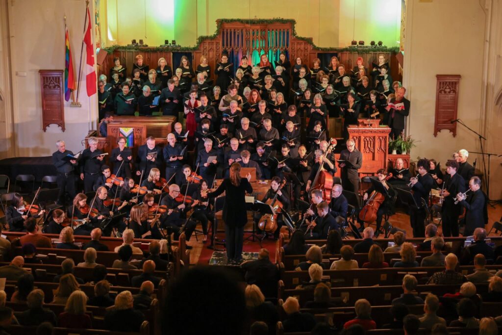 Un immense groupe donne un concert, avec un orchestre au sol et des chanteurs occupant les marches d'une église. Une femme vue de dos dirige l'ensemble.