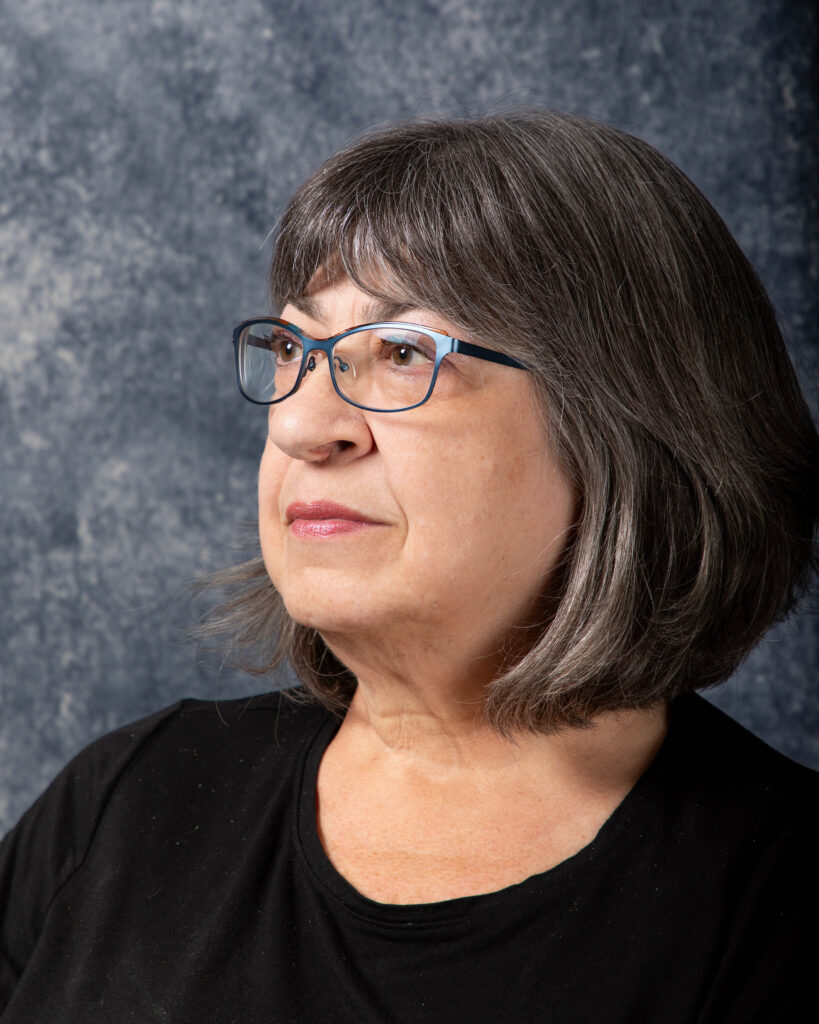 A white woman with medium-length black hair and black glasses, wearing a black top. Her head is slightly turned to the right.