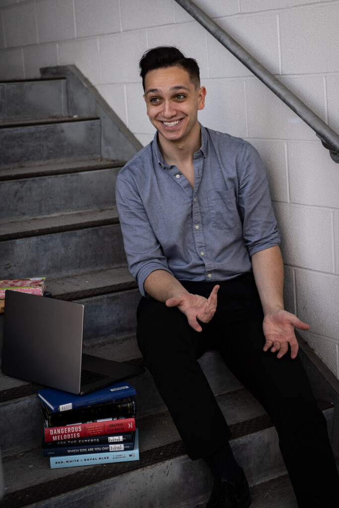 A white man with short black hair wearing a blue shirt and black pants, smiling at the camera, sitting in a grey staircase close to a pile of books with an open laptop on it. The hands seem in movement as if accompanying a vivid conversation.