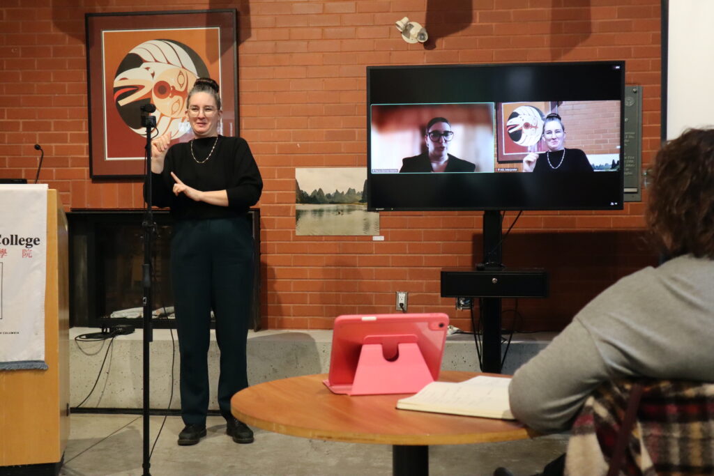 A white woman dressed in black interprets in sign language. The head of a white woman with black glasses presenting is displayed on a TV screen.