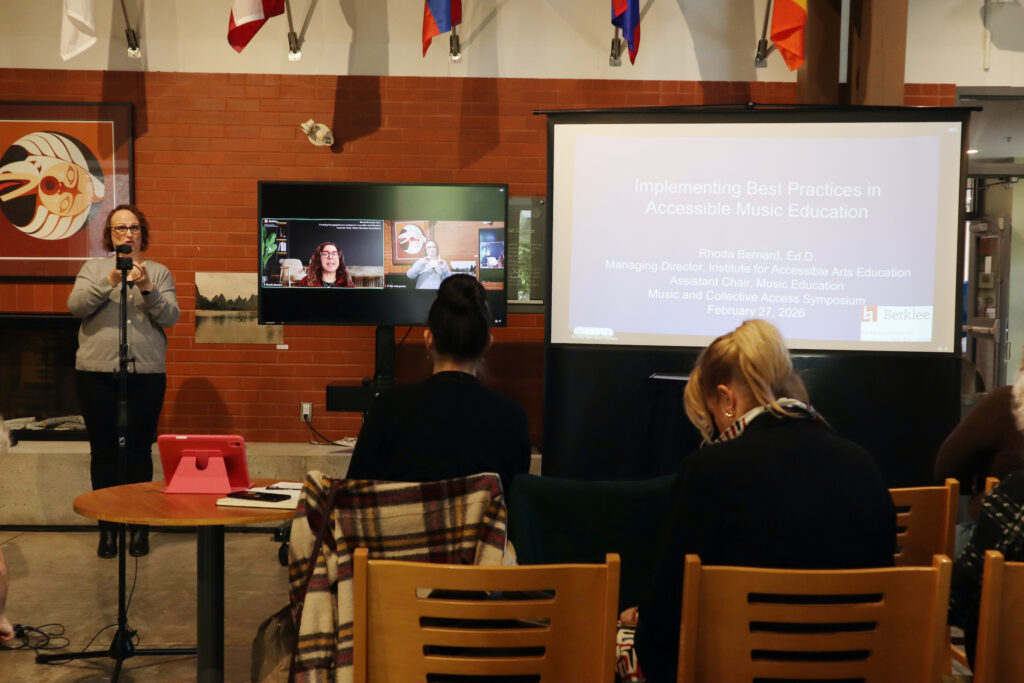 A white woman dressed with glasses and a grey sweat shirt interprets in sign language. The head of a white woman with red glasses presenting is displayed on a TV screen. Her slideshow is projected on a larger screen on the right.