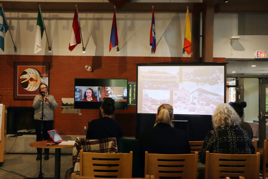 A white man with curly hair and glasses wearing a red top presents. His head appears on the TV screen. Pictures of the desert are displayed on the larger screen on the right.