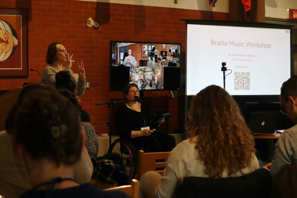 A white woman with glasses wearing a black top sits in a wheelchair and speaks in a microphone. A presentation of a workshop about Braille Music is displayed on the larger screen.
