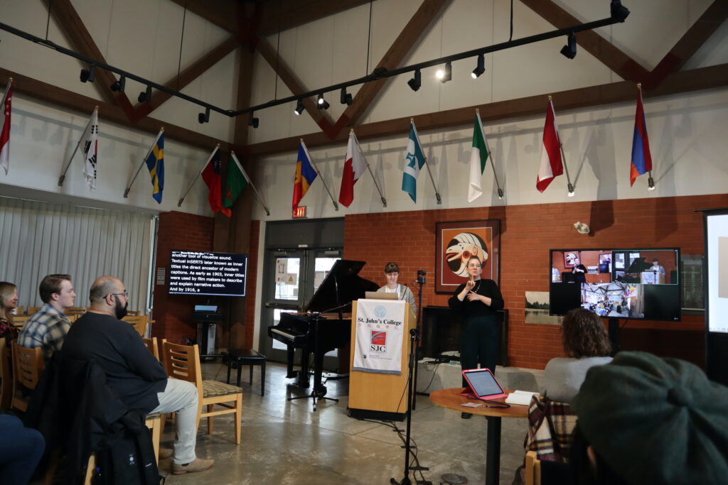A white woman with dark hair presenting from a wooden stand with the St. John's College logo on it.