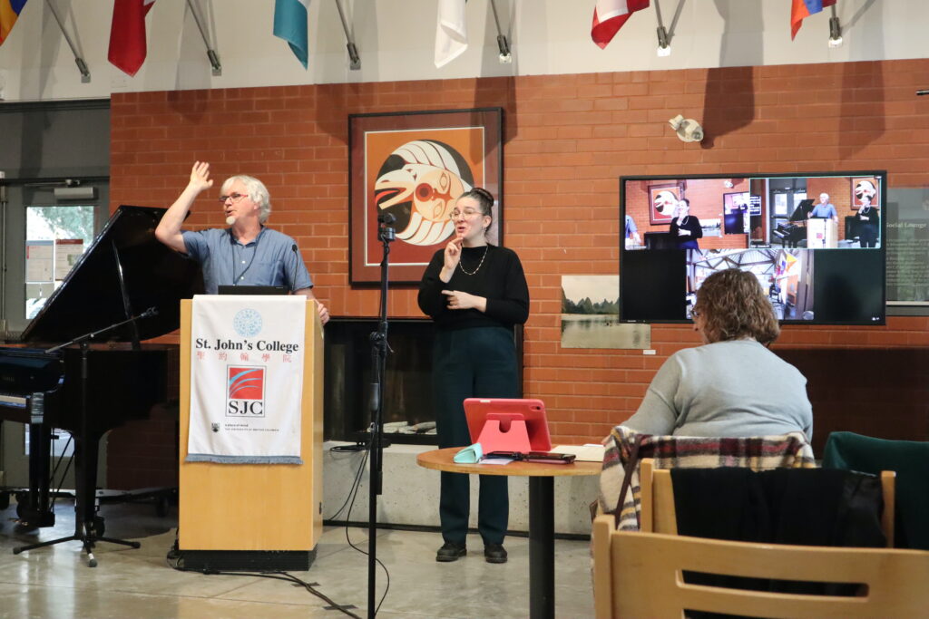 A white man with white hair and glasses wears a blue short. He presents from the wooden stand. His right arm is raised.