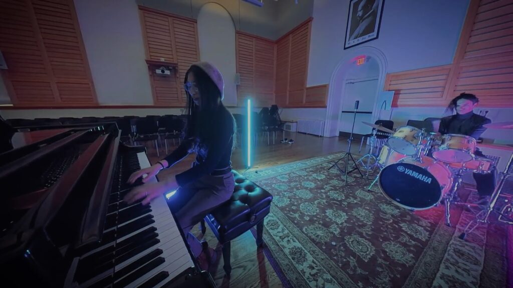 An Asian woman with glasses and long black hair playing the piano on the left, and an Asian man on the right playing the drums. The lights are dimmed, except for a LED in the background.
