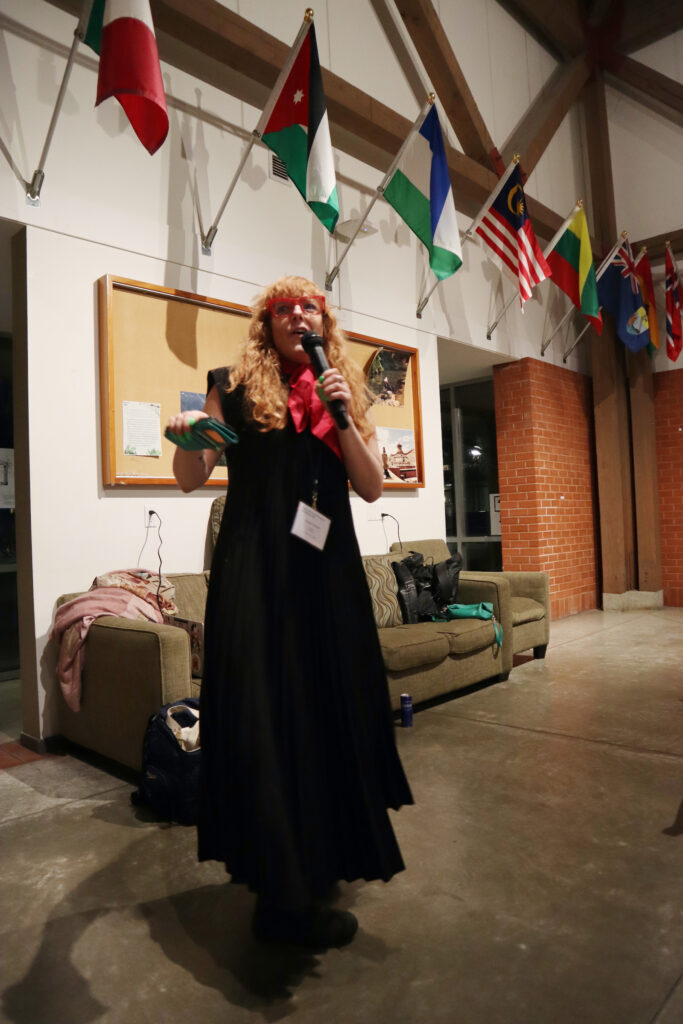 A white woman with red curly hair and red glasses wearing a black dress holds a microphone and gives a speech.