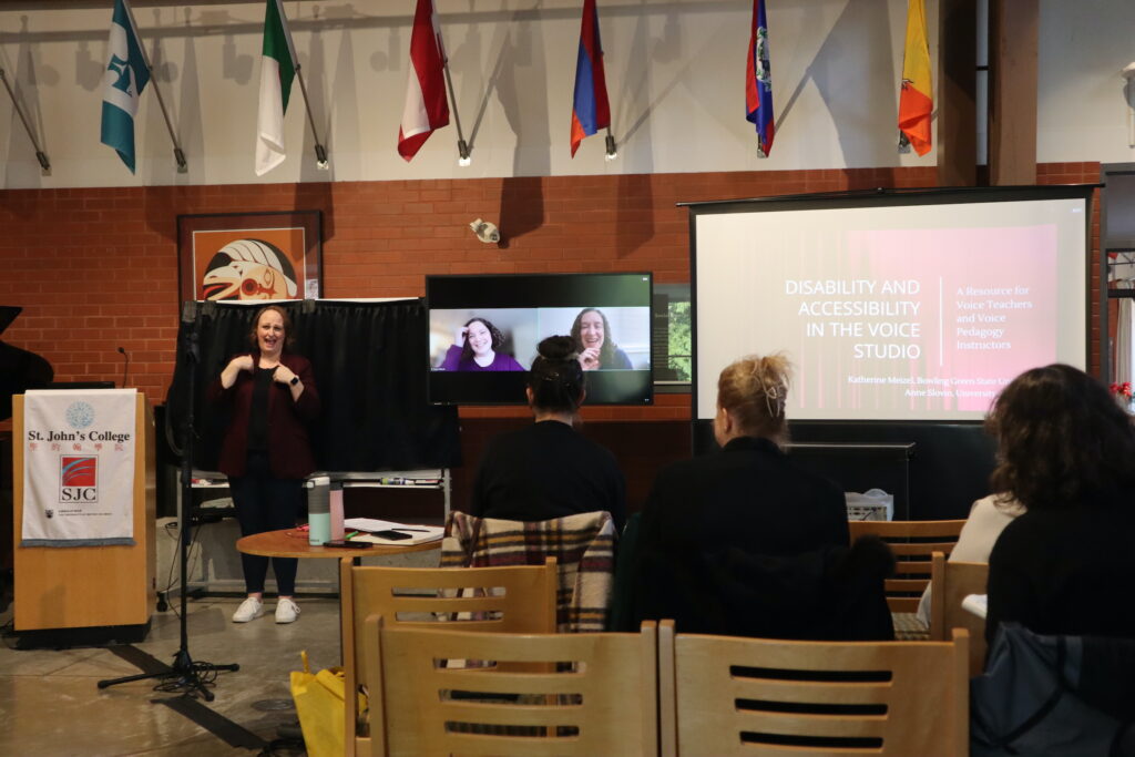 Two white women present remotely, their faces are shown on the screen. Their presentation about accessibility in the voice studio is displayed on the larger screen on the right.