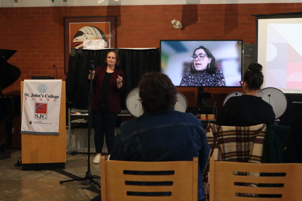 A white woman with dark hair wearing black glasses presents remotely, her face is shown on the TV screen.
