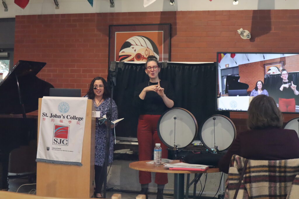 A white woman with glasses wearing a blue dress holds a notebook and speaks from the wooden stand.