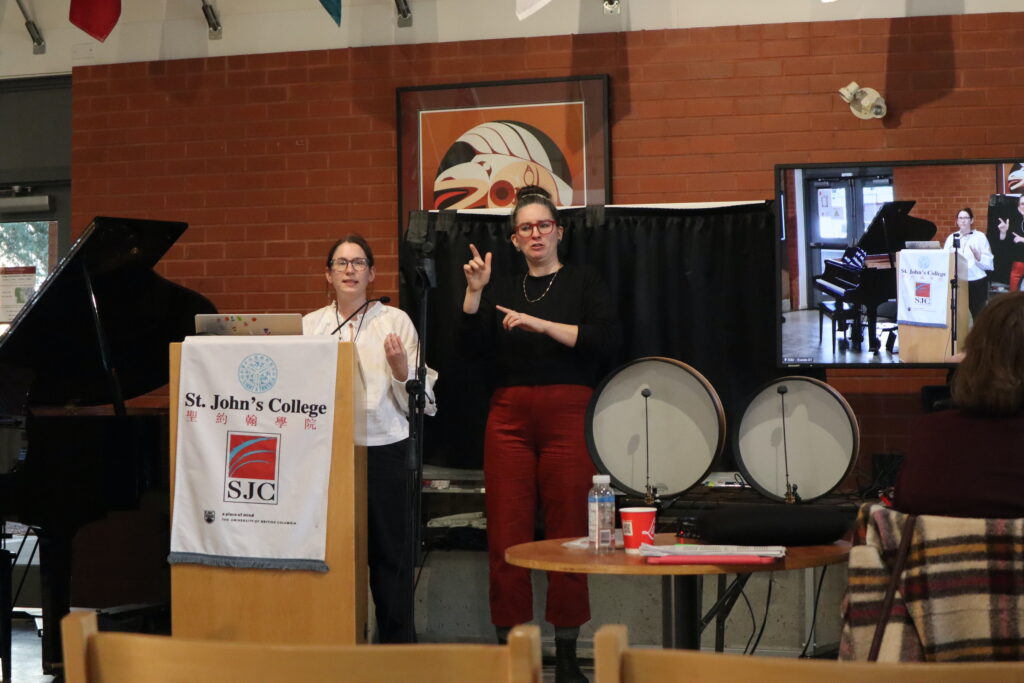 A white woman with brown hair and glasses wearing a white shirt presents from the wooden stand.