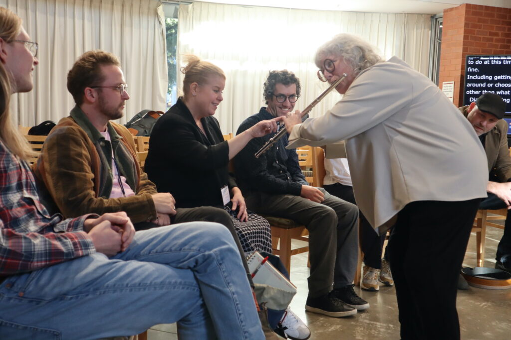 A white woman with white curly hair and black glasses wearing a beige vest and black pants plays the flute, participants feel the vibration of the instrument.