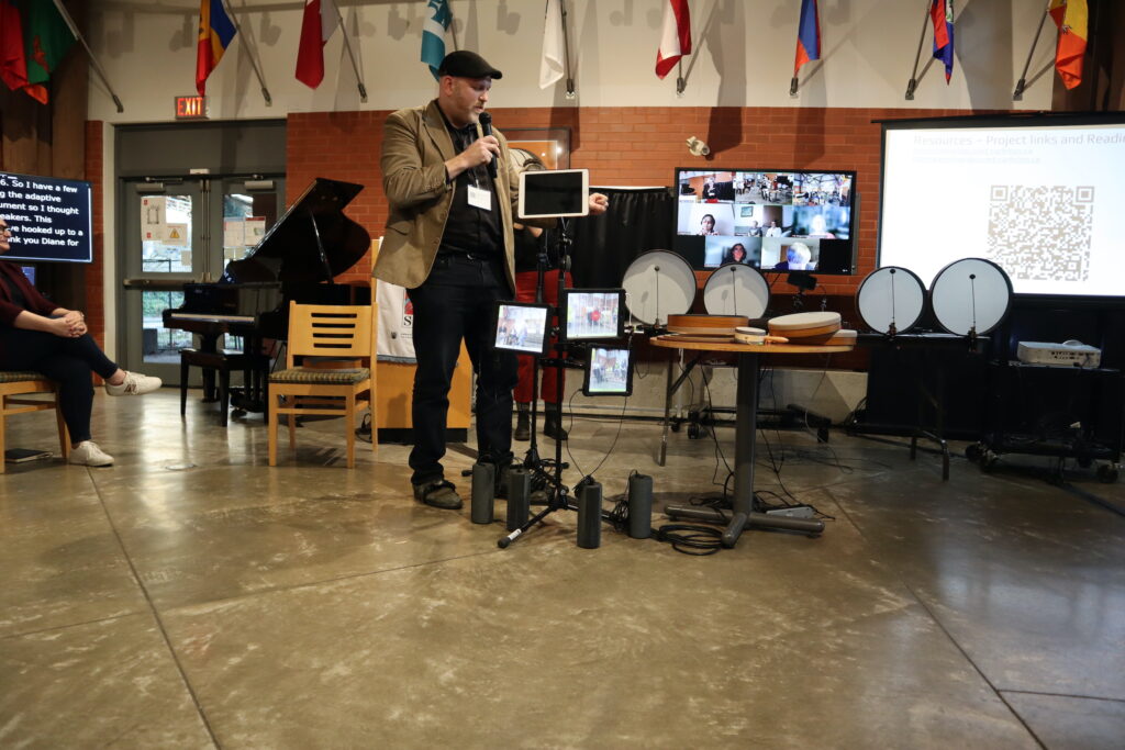 A white man wearing a brown vest on a black top and black pants presents a series of drums and connected instruments on ipads.