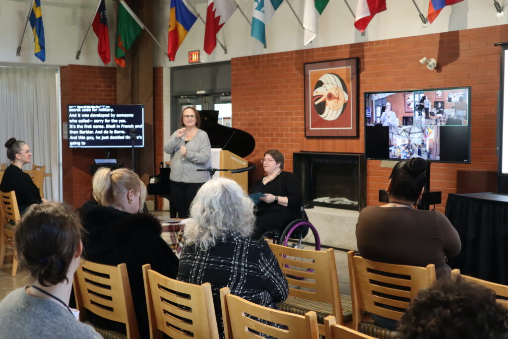 A white woman with glasses wearing a black top sits in a wheelchair and speaks in a microphone, in front of a crowd. A white woman with glasses and brown hair on her left signs in sign language.