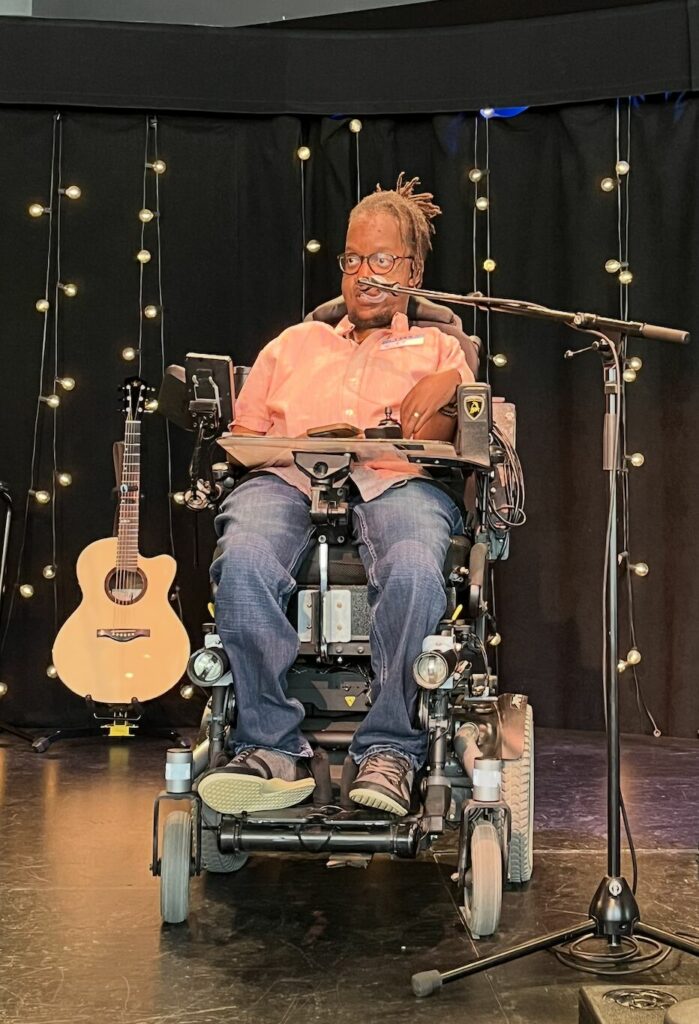 A black man with a pink shirt and blue jeans, wearing black framed glasses, using an electric wheelchair. He talks in a microphone on a stage. Behind him is a guitar.