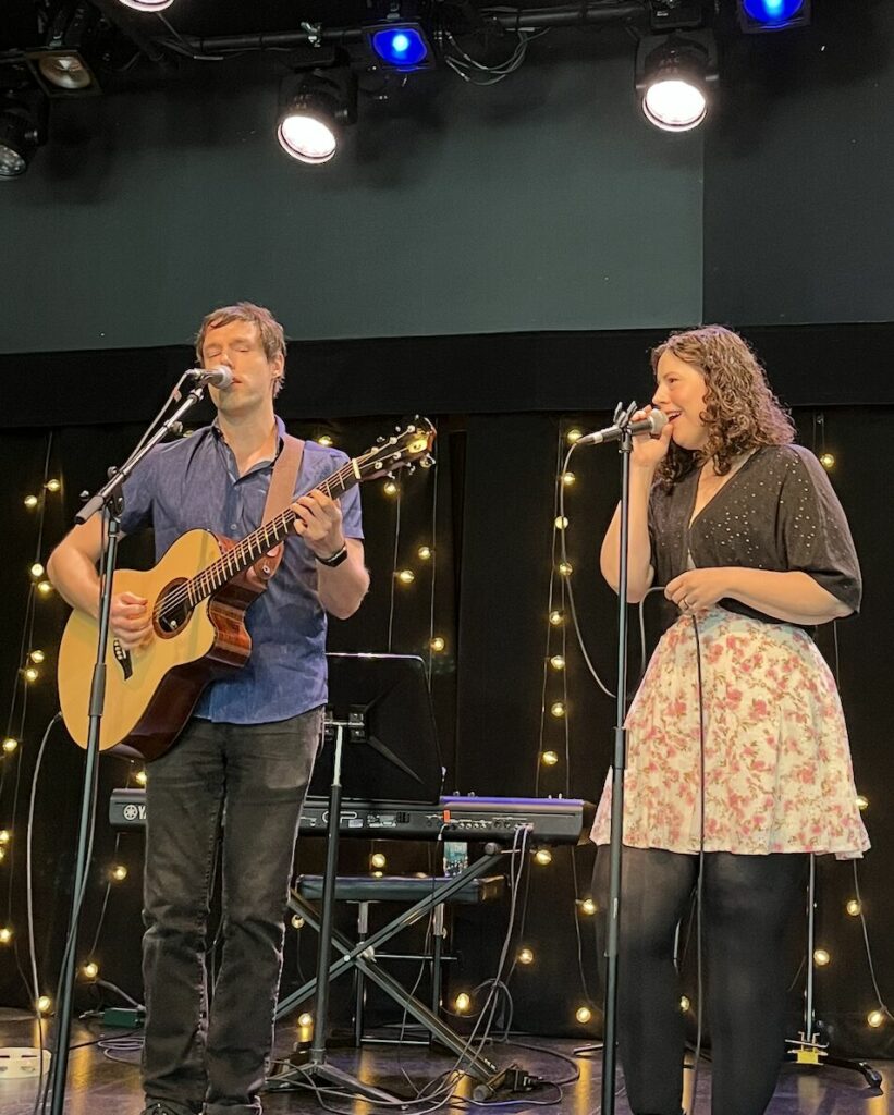 On the left, a white man with short brown hair wearing a blue shirt and black pants holds a guitar and sings in a microphone. On the right, a white woman with long curly brown hair wearing a black top and a pink skirt sing in a microphone.