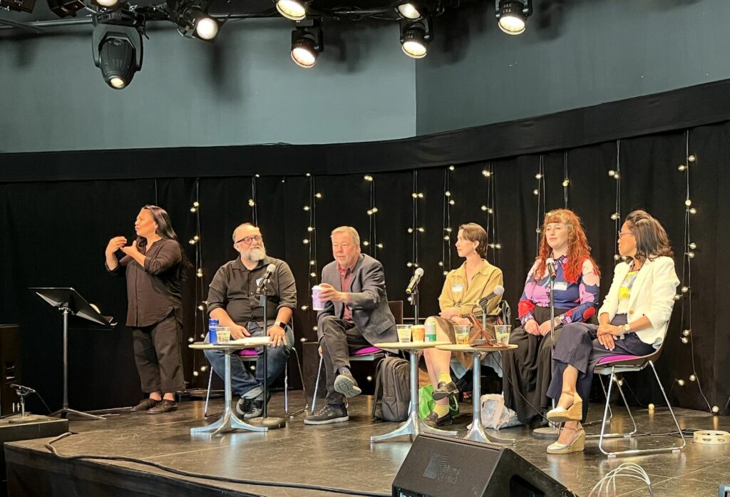 Six individuals on a stage. The one on the left stands and sings in ASL. The five others sit on chairs on a stage.