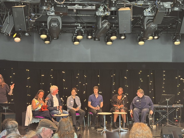 Seven individuals on a stage. The one on the left stands and sings in ASL. The six others sit on chairs on a stage.