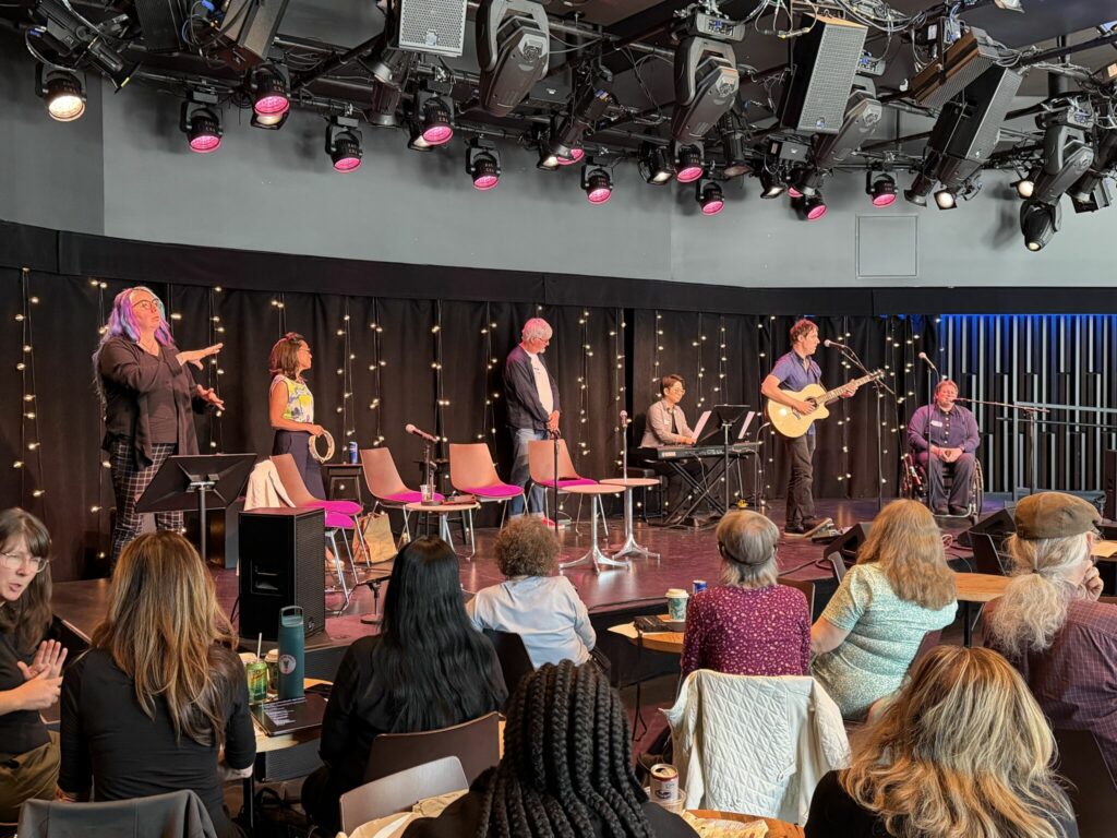 Six individuals on a stage. The one on the left stands and signs in ASL. One person stands with a tambourine in hand. One person stands close the piano. One person sits in front of the piano, playing. One person stands in front of a microphone and plays the guitar. One person sits in a wheelchair in front of a microphone and sings.