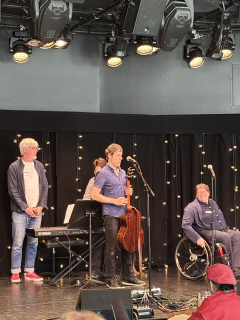 Four persons on a stage, ready to salute after a performance. The two pianists stand close to the piano, the guitarist holds his guitar, and the singer in a wheelchair smiles.