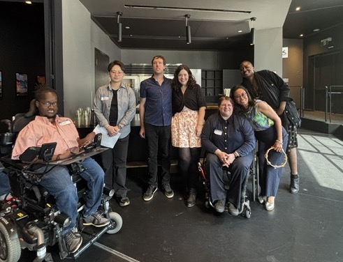 Seven people smiling at the camera for a picture. One uses an electric wheelchair, one a manual wheelchair, and the others stand.